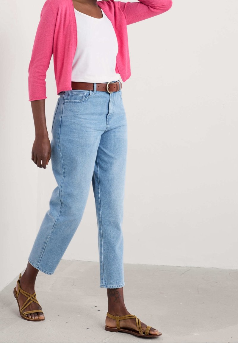 Personne portant un jean bleu clair, une ceinture marron, un haut blanc, un cardigan rose et des sandales marron à brides, debout devant un fond clair uni.