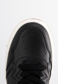 Schwarzer Sportschuh mit Obermaterial aus Leder, perforierter Zehenpartie, weißer Zwischensohle und schwarzen Schnürsenkeln. Verfügt über ein gemustertes Seitenpanel und eine Laufsohle aus Gummi.