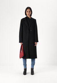 Svart lang kåpe med krage, knappdetaljer, parret med blå jeans og svarte ankelstøvler. Holder en liten rød håndveske.