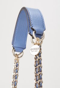 Tracolla in pelle blu con catena dorata e un'etichetta metallica rotonda incisa con "GUESS EST. 1981" attaccata a un anello metallico.
