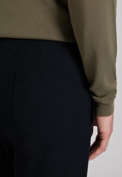 Granatowe spodnie z elastycznym pasem, z tylną naszywaną kieszenią, nad którą widać teksturowany beżowy rękaw swetra.