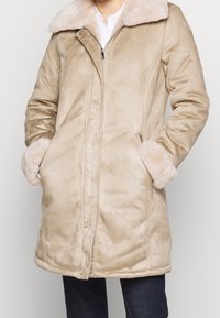 Abrigo beige forrado con piel sintética, con cierre de cremallera, bolsillos laterales y cuello, que presenta una textura suave y un ajuste entallado.