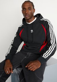 Felpa nera con accenti rossi e strisce bianche lungo le maniche. Presenta un logo sul petto e un cappuccio con cordino. Indossata con jeans scuri.