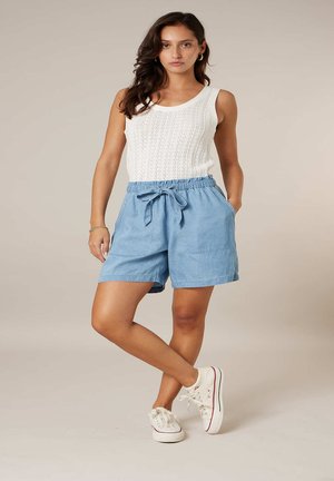 Femme debout, une jambe croisée, portant un haut en maille sans manches blanc, un short bleu clair noué à la taille, des baskets blanches et des boucles d'oreilles créoles dorées.