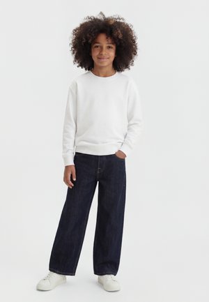 Kind mit lockigem Haar, das ein weißes Langarmshirt, dunkle Jeans mit weitem Bein und weiße Sneakers trägt, steht mit einer Hand in der Tasche.