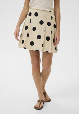 Beiger plissierter Rock mit großen schwarzen Polka-Dots, kombiniert mit einem hellen Tanktop und braunen Zehensandalen bei einer stehenden Person.