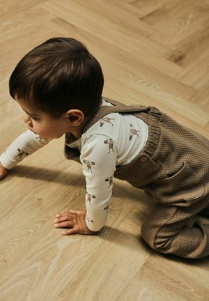 Dziecko w wzorzystej koszulce z długim rękawem i teksturowanych brązowych ogrodniczkach, czołgające się po drewnianej podłodze. Ogrodniczki mają guziki i boczne kieszenie.