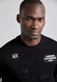 Črna dukserica s vezljivimi logotipi, vključno z "AIX" in "ARMANI EXCHANGE 1991," z okroglo ovratnikom in strukturiranimi detajli.