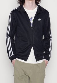 Person med en svart Adidas zip-up jacka med vita ränder på ärmarna, en vit grafisk t-shirt, beige byxor och ett silverarmband.