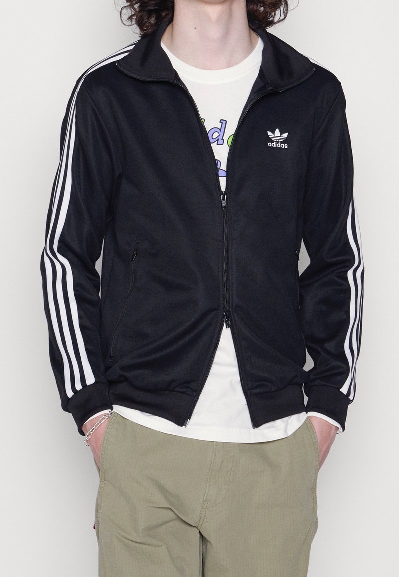 Person med en svart Adidas zip-up jacka med vita ränder på ärmarna, en vit grafisk t-shirt, beige byxor och ett silverarmband.