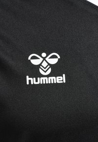 Sort stof med et hvidt logo af en stiliseret bi og brandnavnet "hummel" i moderne typografi. Glat tekstur, atletisk design.