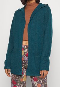 Tealstickad cardigan med huva, öppen front och två framfickor. Bärs över en brun topp och blommönstrade byxor.