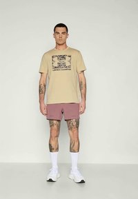 Vyras stovi, vilkėdamas kreminės spalvos "Under Armour" marškinėlius, melsvus šortus, baltas kojines ir baltus sportbačius, ant rankų ir kojų rodo tatuiruotes.