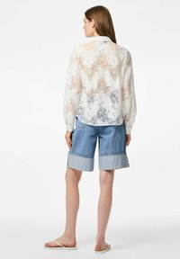 Witte blouse met bloemenpatroon, losse mouwen en een gebogen zoom, gecombineerd met lichtblauwe denim shorts met opgerolde mouwen.