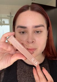 ILIA True Skin Serum Concealer i ett ljusrosa rör med vit text; produkten hålls i en hand mot en neutral bakgrund.