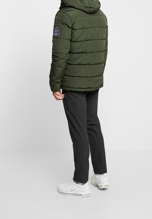 Persona con una chaqueta de plumas verde oscuro con un parche de logo en la manga, pantalones gris oscuro y zapatillas Nike blancas, de pie sobre un suelo blanco.