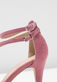 Zapato de tacón alto de ante rosa con dobles correas en el tobillo aseguradas por hebillas plateadas. Forro interior suave y diseño de tacón estructurado.