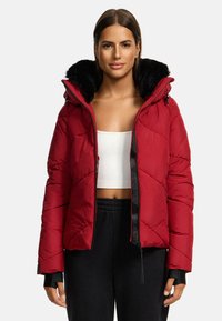 Doudoune rouge avec capuche en fausse fourrure, texture matelassée, fermeture éclair à l'avant et accents noirs. Associée à un crop top blanc et un pantalon noir.