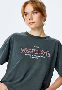 Szary t-shirt z grafiką z wyraźnym napisem "DISCHARGE" w czerwonych konturowych literach, z dodatkowym białym tekstem poniżej. Krótkie rękawy, luźny krój.