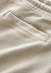 Beige szövet sima textúrával, oldalán részes zsebbeállással, varrott szélekkel és enyhe nyújthatósággal.