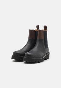Botas Chelsea de cuero negro con acentos marrones, paneles laterales elásticos y una suela de goma gruesa con clavos pronunciados para tracción.