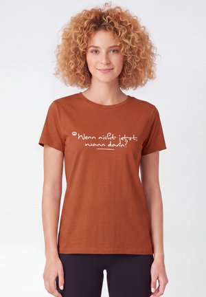 Frau mit lockigem Haar, die ein rostfarbenes T-Shirt mit deutschem Text "Wenn nicht jetzt, wann dann?" vor einfachem weißem Hintergrund trägt.