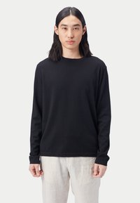 Schwarzes langärmeliges Hemd in entspannter Passform, aus weichem Stoff mit Rundhalsausschnitt. Kombiniert mit hellen Leinenhosen. Schlichtes Design, ohne Muster.