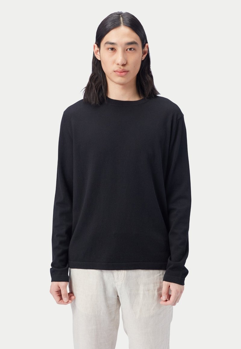 Schwarzes langärmeliges Hemd in entspannter Passform, aus weichem Stoff mit Rundhalsausschnitt. Kombiniert mit hellen Leinenhosen. Schlichtes Design, ohne Muster.