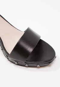 Sandale à glissière noire avec une large bride lisse, une semelle plate et des clous métalliques le long du bord pour un détail supplémentaire.