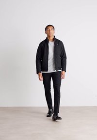 Zwarte gewatteerde jas met een staande kraag, gedragen over een grijs T-shirt. Slanke zwarte broek en zwarte schoenen maken de look compleet. Eenvoudige achtergrond.