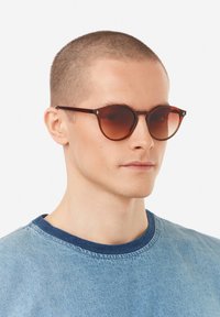 Pyöreät, ruskehtavat aurinkolasit, joissa on ohut, tummanruskea kehys. Hienovaraiset logoaktsentit temppeleissä. Yhdistetty siniseen pyöreäkaulaiseen paitaan.