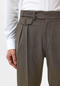 Bruine gestreepte broek met een geplooid ontwerp, voorzien van een knoopsluiting en riemlussen. De stof heeft een gestructureerde afwerking.