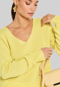 Helle gelbe Strickjacke mit V-Ausschnitt und langen Ärmeln, kombiniert mit einer beigen Handtasche. Weiche Textur, minimalistisches Design, keine Muster.
