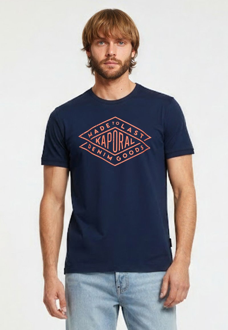 Homme portant un t-shirt bleu marine avec le logo orange "Kaporal Made to Last Denim Goods", associé à un jean bleu clair, debout devant un fond uni.