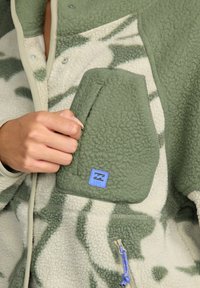 Zielona i kremowa polarowa kurtka w wzór kamuflażowy; posiada kieszeń z przodu oraz niebieską naszywkę z logo; teksturowana tkanina i zamki błyskawiczne.