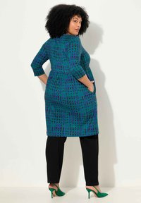 Türkis-violettes gemustertes Kleid mit 3/4-Ärmeln und Taschen, kombiniert mit schwarzen Hosen und grünen High-Heels.