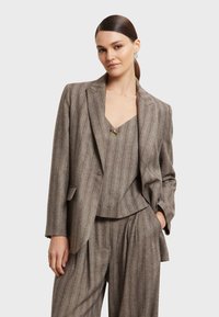 Grijze geruite pak gemaakt van gestructureerde stof, met een op maat gemaakte blazer met gegolfde revers en geplooide, wijde broekspijpen. Accenten in goudkleurig metaal.