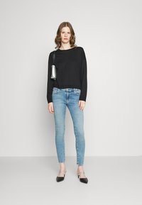 Svart langermet topp, lyseblå skinny jeans, og svarte spissede slingback sko. Liten sølvbag på skulderen. Enkel bakgrunn.