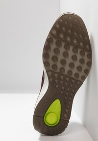 Semelle d'une chaussure marron et blanche avec une surface antidérapante texturée, des crampons circulaires et un insert circulaire jaune-vert près du talon.