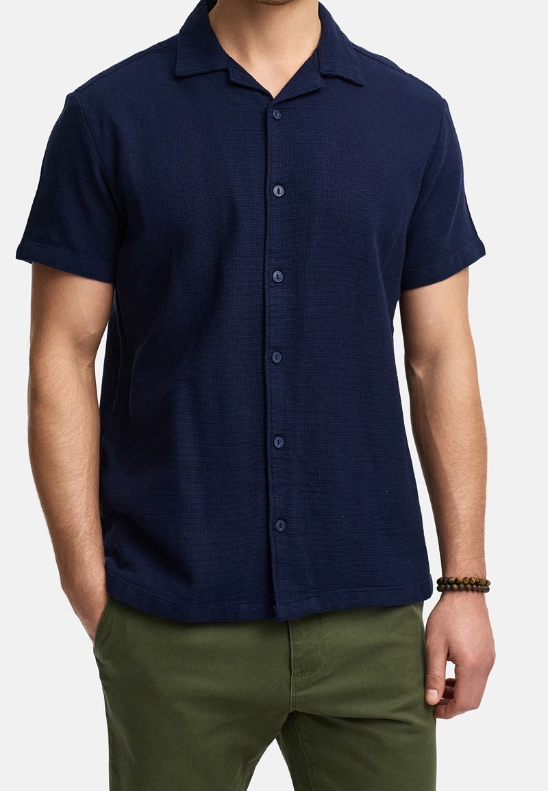 Uomo che indossa una camicia blu navy a maniche corte con colletto, pantaloni verde oliva e un braccialetto di perline al polso destro.