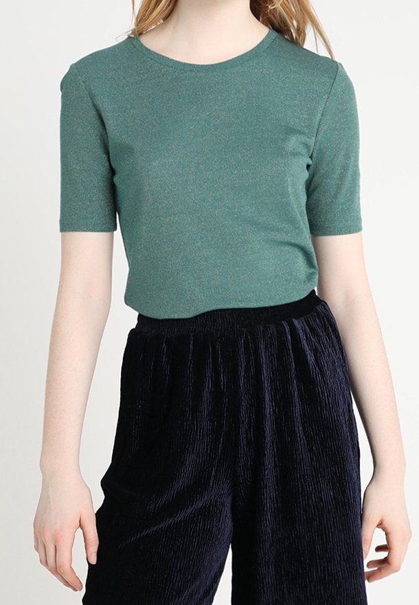 Femme portant un haut vert ajusté à manches courtes et un pantalon noir texturé, taille haute et jambes larges, sur un fond uni.