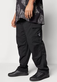 Mies, joka pitää yllä mustia cargo-housuja Calvin Klein -logopatchilla, mustia lenkkitossuja ja mustaa kuvioitua paitaa, seisoo tasaisen taustan edessä.