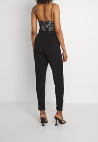 Svart jumpsuit med en åtsittande bodice i spets och tunna axelband som övergår till vida, avsmalnande byxor. Har en slät yta och minimal hårdvara.