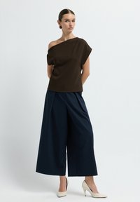 Top scuro marrone a spalle scoperte abbinato a pantaloni larghi navy. L'outfit presenta texture morbide e tacchi alti color crema.