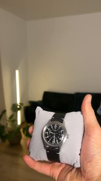 Montre Timex avec cadran noir, boîtier en acier inoxydable argenté et bracelet en cuir noir. La montre est présentée sur un coussin gris.