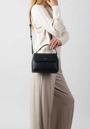 Mujer vestida con suéter color crema y pantalones beige, sosteniendo un bolso de cuero negro con asa superior y correa para el hombro sobre un fondo claro.