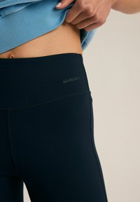 Czarne legginsy sportowe z wysokim stanem, charakteryzujące się gładką teksturą i subtelnym brandingiem w talii. Projekt ma dopasowany krój.
