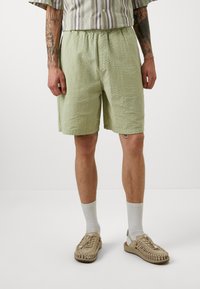 Bermudas verde claro y texturizadas con una cinturilla elástica, por encima de la rodilla. Combinadas con calcetines crema y sandalias trenzadas color tostado.