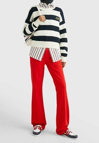Gestreifter Marineblauer und Cremefarbener Pullover mit geripptem Saum, über einem rot-weißen gestreiften Hemd getragen. Rote Schlaghosen und marineblaue Schuhe mit weißen Akzenten.
