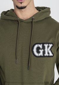 Felpa verde oliva realizzata in tessuto morbido, con un logo ricamato nero "GK". Include cordini e una tasca a marsupio.